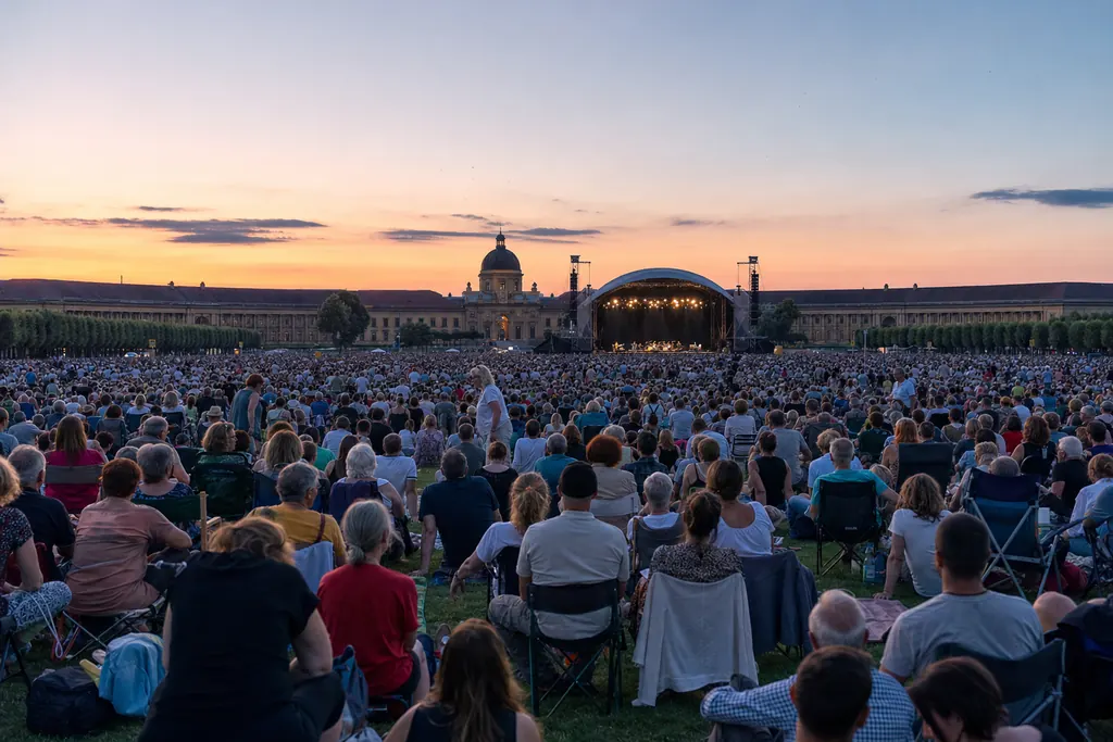 Pubblico di un concerto all'aperto sul prato al tramonto davanti a un grande palco illuminato (immagine creata con AI)