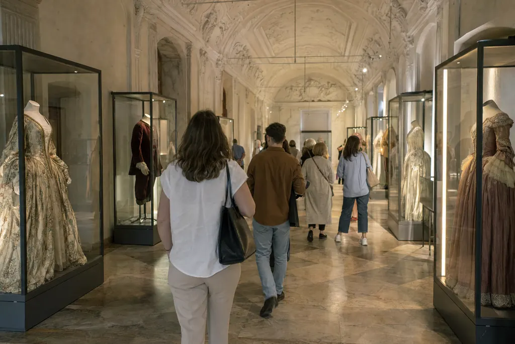 Visitori di spalle in una lunga sala barocca allestita con costumi storici in vetrine (immagine creata con AI)